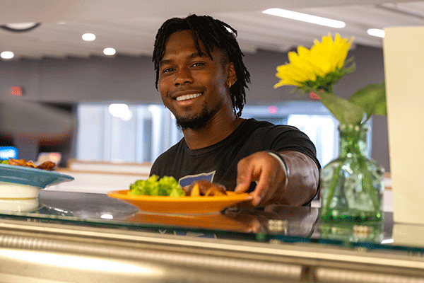 A Black male student smiles as he is handed a plate in line at the Sycamore Dining Hall.  