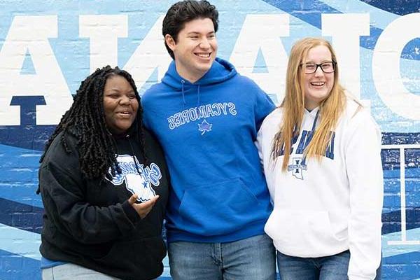 Three students standing in front of a blue-and-white painted Indiana State mural. On the far left is a white female student with straight blonde hair. She wears a white Indiana State sweatshirt, black glasses, and blue jeans. In the middle is a white male student with black hair. He wears a blue Sycamores sweatshirt and blue jeans. On the right is a Black female student with black dreadlocks. She wears a black Sycamores sweatshirt and blue jeans.  